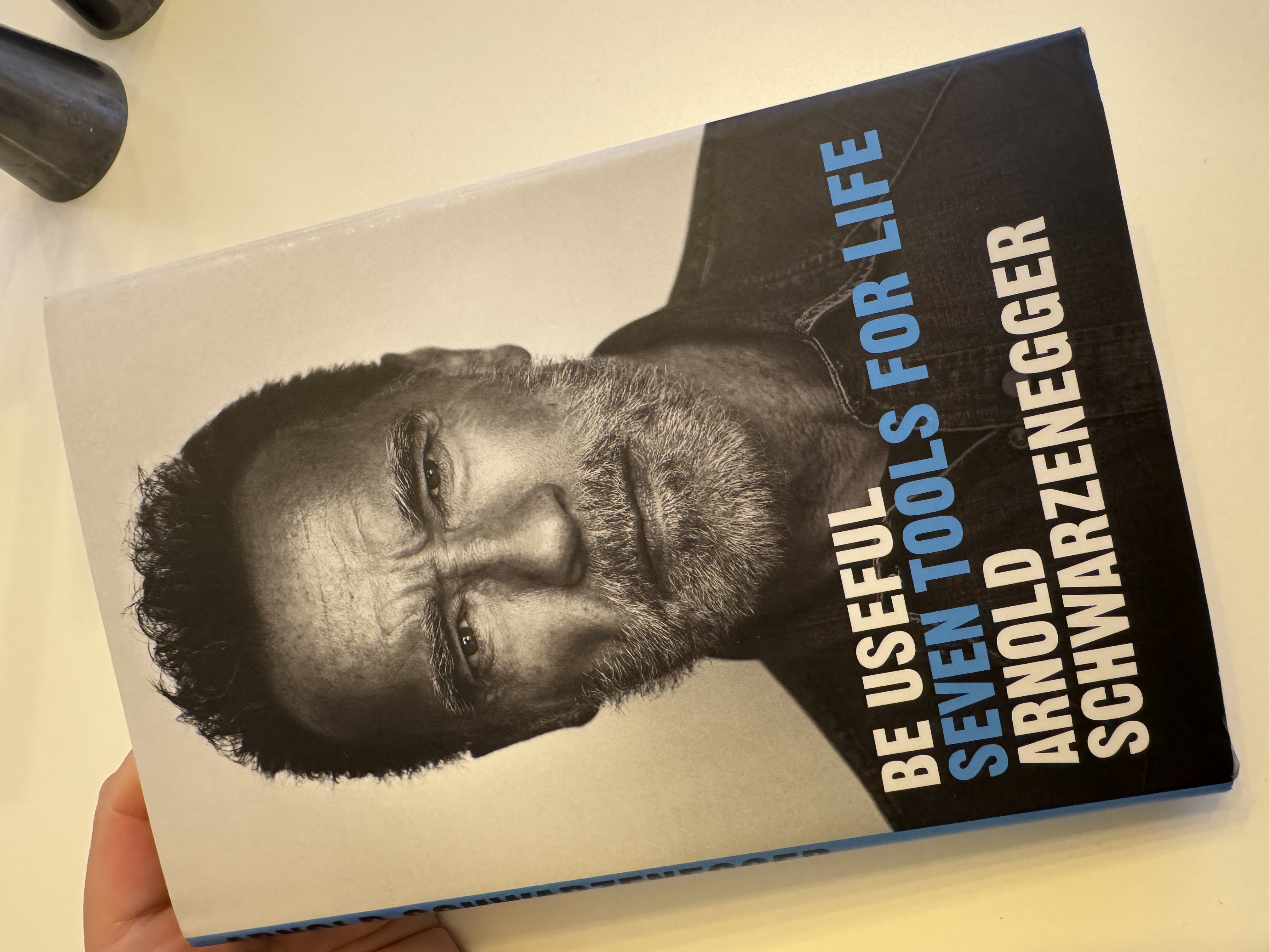 Book: Be Useful – Seven Tools for Life, by Arnold Schwarzenegger.
The cover is a black-and white photo portrait of Schwarzenegger - now 76 years old - looking determined.
The book is on my white breakfast table and I'm supporting the top of it with my left hand.