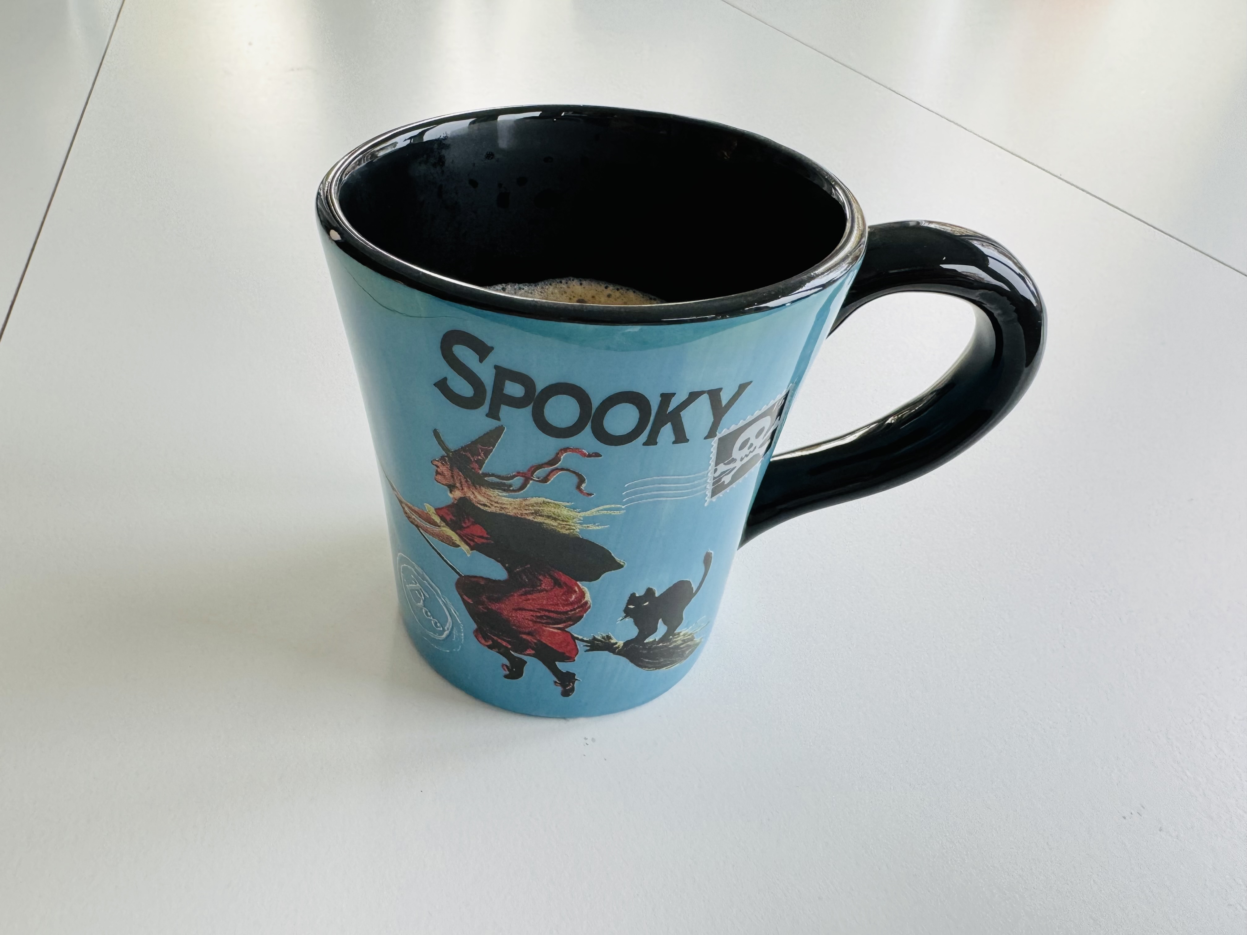 A blue mug with an illustration of a witch flying on a broom and a black cat riding with them. There is also a postage stanp with a skull like Jolly Roger, and the word Spooky is written above the scene.

In the mug is coffee.