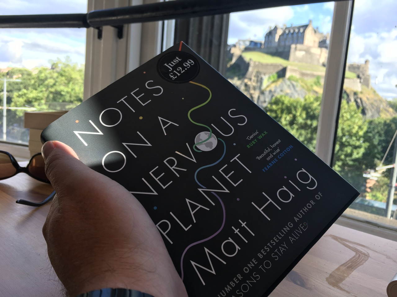 Hand holding the book Notes on a Nervous Planet by Matt Haig. Through the window in the background one can see Edinburgh Castle.