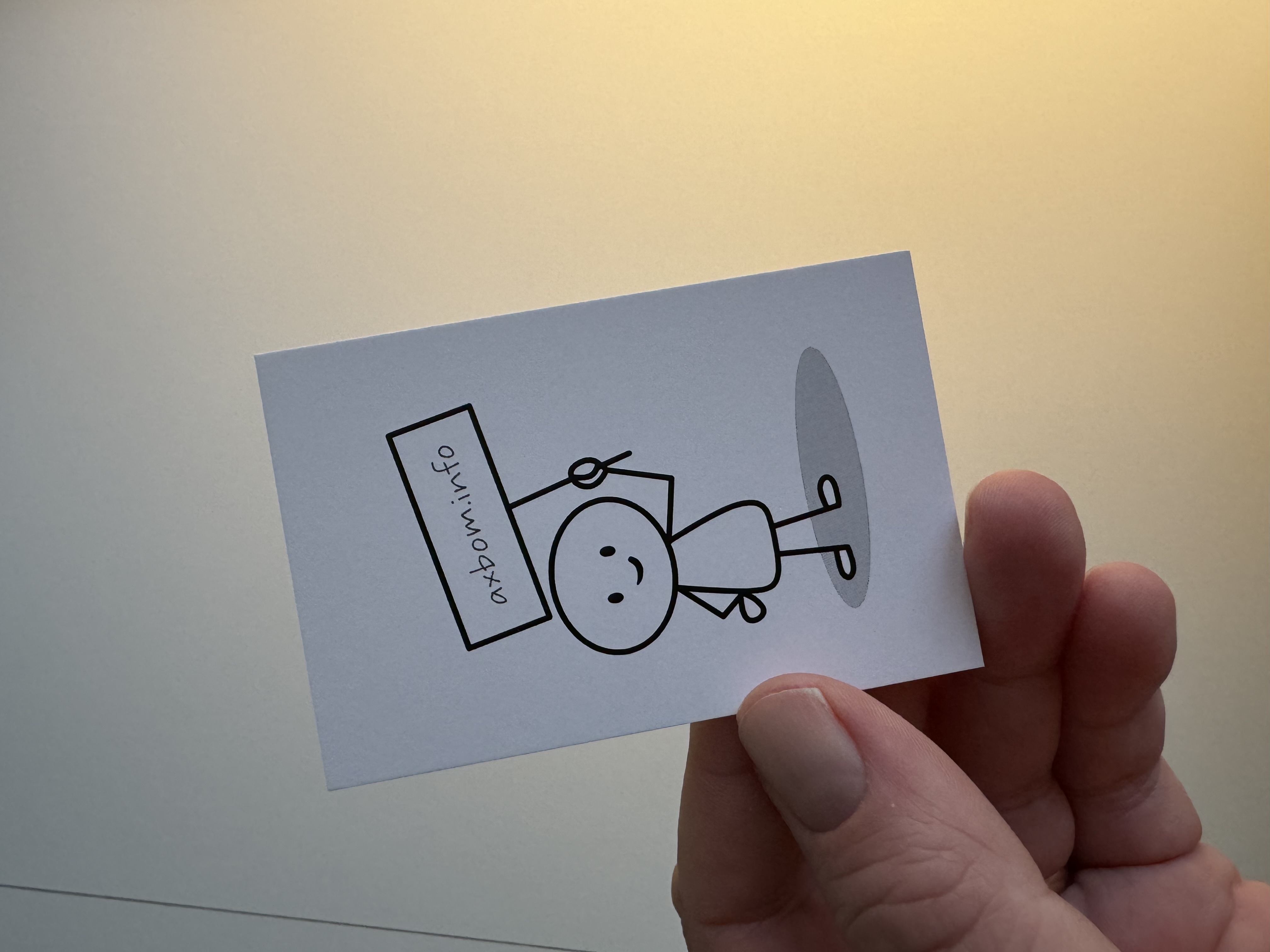 Hand holding a business card with a simple line drawing of a person holding a sign. The sign reads