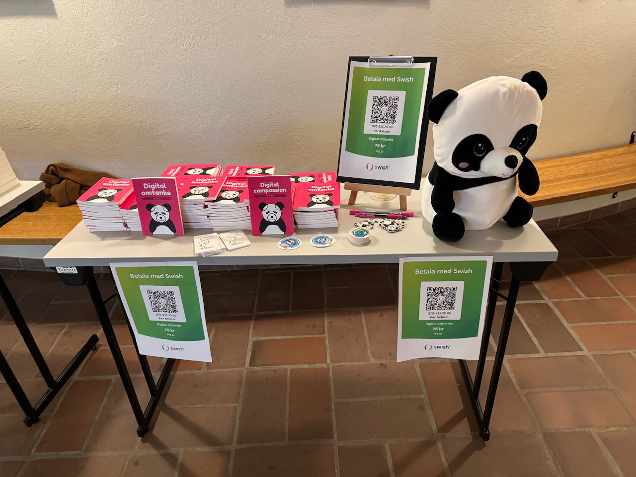 Bord med flera staplar med min bok Digital omtanke (med en panda på omslaget), klistermärken, affischer för att betala med Swish, och ett stort panda-gosedjur.